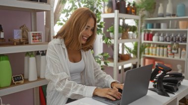 Ev dekorasyonu mağazasında dizüstü bilgisayarda çalışan genç bir kadın. Etrafı çeşitli dekoratif ürünler ve bitkilerle çevrili.