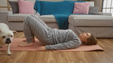 Genç sarışın kadın oturma odasında yoga minderinde egzersiz yapıyor. Yanında da bir köpek var..