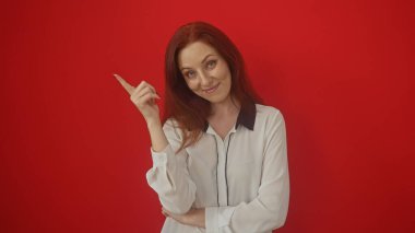 Beyaz bluzlu, kendine güvenen genç, kızıl saçlı bir kadın sağlam kırmızı bir arka planda hareketlenir, büyüleyici ve pozitiftir..