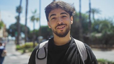 Arkasında palmiye ağaçları olan güneşli bir şehir sokağında gülümseyen sakallı yakışıklı bir Arap..