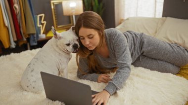 Genç sarışın bir kadın, yatak odasında evcil köpeğiyle birlikte bir yatakta uzanıyor. İkisi de laptopa bakıyor. Kapalı alanda rahat bir sahne yaratıyor..