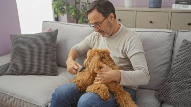 Orta yaşlı bir adam kanepede kanişi tımar ediyor, evdeki rahat bir evcil hayvan bakımını tasvir ediyor..