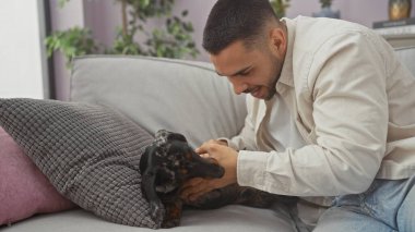 Oturma odasındaki kanepede köpekle oynayan gülümseyen genç adam..