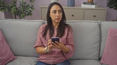 Genç bir kadın evdeki bir kanepede oturmuş, elinde akıllı bir telefonla endişeli endişeli bakıyor..