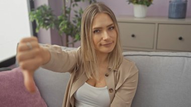 A beautiful blonde woman in a living room shows a displeased thumbs-down gesture while sitting on a couch in a cozy home interior. clipart