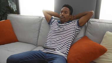 Modern bir oturma odasındaki rahat bir kanepede dinlenen genç bir adam, şık bir iç tasarım ile evin içinde sakin ve rahat bir atmosfer sergiliyor..