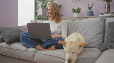 Rahat bir oturma odasında uyuyan köpeğinin yanında dizüstü bilgisayar kullanan genç bir kadın..