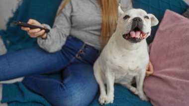 Bir elinde televizyon kumandası tutarken elinde mutlu bir köpekle oturma odasında uzanan genç bir kadın.