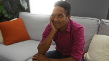 Kapalı bir kanepede oturan genç adam, elleri çenesinde, kırmızı bir gömlek giyiyor, rahat bir oturma odasında, rahatlama ve tefekkür düşüncelerini aktarıyor..