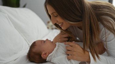 Anne ve bebek sıcak yatak odası ortamında yatakta sevgi dolu bir şekilde kaynaşıyor, aile sıcaklığının ve birlikteliğin özünü yakalıyorlar..