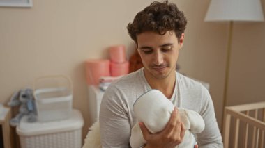 Genç adam sıcak yatak odasında bebeği tutuyor, şefkat ve sıcaklık gösteriyor, sıcak bir beşiğin altında, temiz bir iç mekanda.