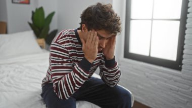 Yatak odasındaki yatakta oturan genç adam derin düşüncelere dalmış, el ele tutuşmuş, ev ortamında stres ya da yorgunluk hissi uyandırıyor..