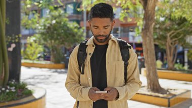 Güneşli bir şehir parkında akıllı telefon kullanan sakallı Afrikalı genç bir Amerikalı..