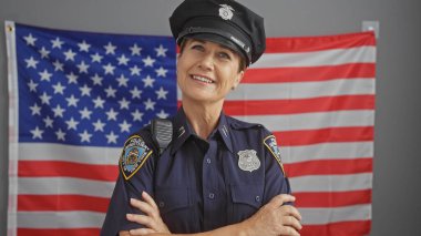 Smiling mature woman police officer with crossed arms in front of american flag indoors. clipart