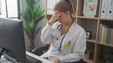 Steteskop, bilgisayar ve yorgunlukla klinikteki stresli sarışın kadın doktor.