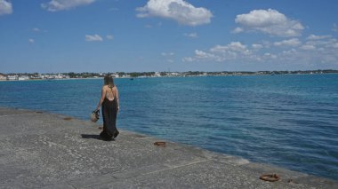 Siyah elbiseli güzel bir İspanyol kadın porto sezeryan, salento, temiz mavi su ve parlak bir gökyüzü ile rıhtım boyunca yürür..