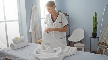 Kadın, yumuşak havlular ve sakin bir atmosferi olan, modern bir klinik ortamında profesyonelliği somutlaştıran ve sakinleştiren parlak bir terapi odasında masaj masası hazırlıyor..