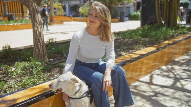 Güneşli bir şehir parkında köpeğiyle bankta oturan gülümseyen genç bir kadın..
