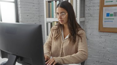 Modern ofis ortamında bilgisayar başında çalışan bir kadın kitap raflarıyla tuğla duvar odaklı ifade ve profesyonel atmosferi aktaran gözlüklerle.