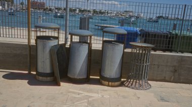 Trash cans along a waterfront in porto cesareo, italy, with boats and buildings in the background on a sunny day. clipart