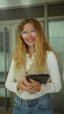 Modern ofiste tabletle gülümseyen bir kadın gözlük takıyor ve teknoloji dostu çalışma alanında doğal ışık ve çağdaş tasarım ile günlük kot pantolon tutuyor..