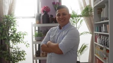 Genç adam, çeşitli eşyalar ve bitkilerle dekore edilmiş iyi ışıklandırılmış bir iç mekan dükkanında güvenle gülümsüyor.