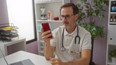 Gözlüklü İspanyol bir adam, klinikte akıllı telefona gülümsüyor, steteskop takıyor, bitkiler ve tıbbi dekor ile çevrili, modern sağlık hizmetleri iletişimini gösteriyor..