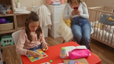 Anne evde rahat bir oyun odasında telefon kullanırken, kız yapboz oynuyor. Renkli oyuncaklar ve sıcak dekor içerikli ev içi aile etkileşimlerini tasvir ediyor..