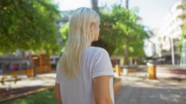 Güneşli bir günde arka planda yeşil ağaçlar ve binalarla şehir parkında yürüyen bir kadın.