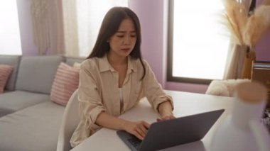 Oturma odasında hapşıran, dizüstü bilgisayar kullanan, bej gömlekli, gri koltuklu, pembe yastıklı, ve masanın üzerine kitap koyan bir kadın.