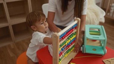 Kadın, oğluyla oyuncak ve beşiği olan rahat bir anaokulunda abaküs kullanarak oynuyor. Ailesini ön plana çıkarıyor ve evde öğreniyor..