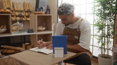 Not defterine sakallı genç adam raflarda ekmek ve hamur işleri sergilenen rahat bir fırında çalışırken bilgisayar tabletine bakıyor.