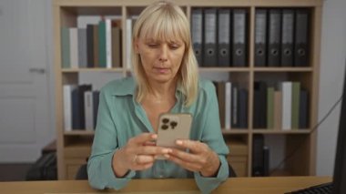 Ofiste akıllı telefon kullanan orta yaşlı bir kadın çalışma masasında iş iletişimi için arka planda raflar olan bir ortam hazırlıyor.