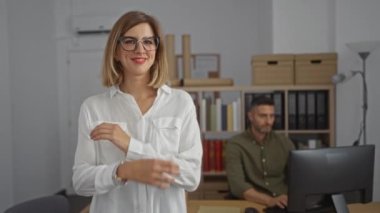 Ofiste kolları çapraz duran ve arka planda bilgisayarın başında çalışan bir adamla gülümseyen kendinden emin bir kadın.