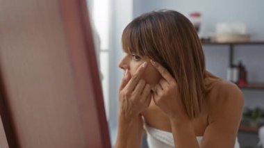 Bir kadın kaplıcada derisini inceliyor, sivilcesine odaklanıyor, arka planda rafları olan bir sağlık odasında..