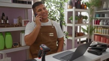 Genç İspanyol bir adam, çeşitli ürün ve bitkilerle dolu kapalı bir dükkânda dizüstü bilgisayarını yönetirken telefonla konuşuyor.