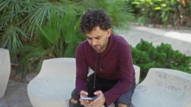 Parktaki bankta oturan genç adam güneşli bir günde arka planında yeşillik olan şehir ortamında akıllı telefonunu kullanıyor..