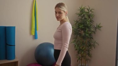 Spor salonunda kollarını kavuşturmuş, spor aletleri ve bitkilerle çevrili bir kadın kendinden emin bir ifade sergiliyor..