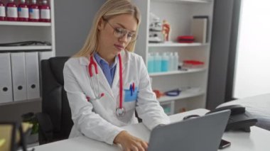 Sarı saçlı ve gözlüklü kadın doktor hastane odasında oturmuş dizüstü bilgisayara bakarken boynuna dokunuyor. Rahatsızlığını gösteriyor..