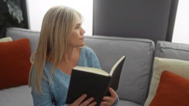 Oturma odasındaki rahat koltukta kitap okuyan bir kadın. Yastıkları ve yumuşak ışıklandırması olan, sarı saçlı, huzurlu bir apartman dairesinde rahat havasını vurgulayan bir kadın..