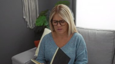 Samimi mavi kazaklı sarışın kadın gözlüklerini çıkarıyor, gözlerini ovalıyor, modern dekorlu ve yakındaki bir pencereden gelen doğal ışıkla şık bir oturma odasında gri bir koltukta oturuyor..