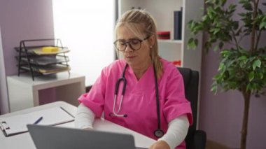 Pembe üniformalı kadın doktor, kliniğin içinde steteskop ve dizüstü bilgisayarla endişeli görünüyor..