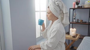 Bir kadın spa odasında dinleniyor, bornoz giyiyor, bardaktan içiyor, mumlar, ürünler ve havlularla çevrili, dingin bir sağlık ortamı sergiliyor.
