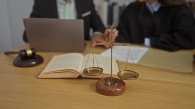Ofisin içinde, yargıç cüppeli bir kadın ve takım elbiseli bir adam yasal belgeleri gözden geçirir. Elinde kitap, terazi ve tahta masada belirgin bir şekilde sergilenen bir tokmak vardır..