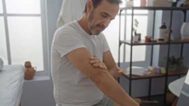 Sakin bir spa ortamında losyon süren olgun bir adam. Yumuşak ışıklandırma ve sağIık dekoruyla. Huzurlu bir ortamda özgüven ve rahatlığı vurguluyor.