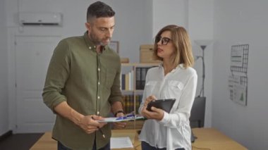 Kadın ve erkek bir ofiste iş birliği yapıyor, çalışma ortamında tablet ve pano üzerinde çalışıyorlar.