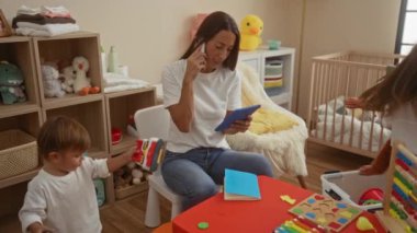 Yakınlarda oynayan bir çocukla aynı anda çocuk yuvasında telefon ve tabletle çalışan kadın, oyuncaklar, beşik ve modern dekor sunuyor, aile hayatını ve ev içi ebeveynlik dinamiklerini ön plana çıkarıyor.