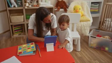 Eğitici oyuncaklar ve canlı dekorlarla çevrili renkli anaokulu odasında tabletli bir çocuğa öğretmenlik yapan bir kadın, kapalı alanda eğlenceli ve ilgi çekici bir öğrenme ortamı yaratıyor..