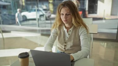 Kafe telefonunda dizüstü bilgisayarda çalışan kadın ön plandaki kahve fincanı ve arka plandaki büyük pencerelerle ilgili gibi görünüyor.