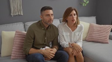 Modern bir oturma odasında gri bir koltukta oturan erkek ve kadın, arka planda pastel yastıklar ve duvar dekorlarıyla bir futbol topunu tutarken hayal kırıklıklarını ifade ediyorlar.